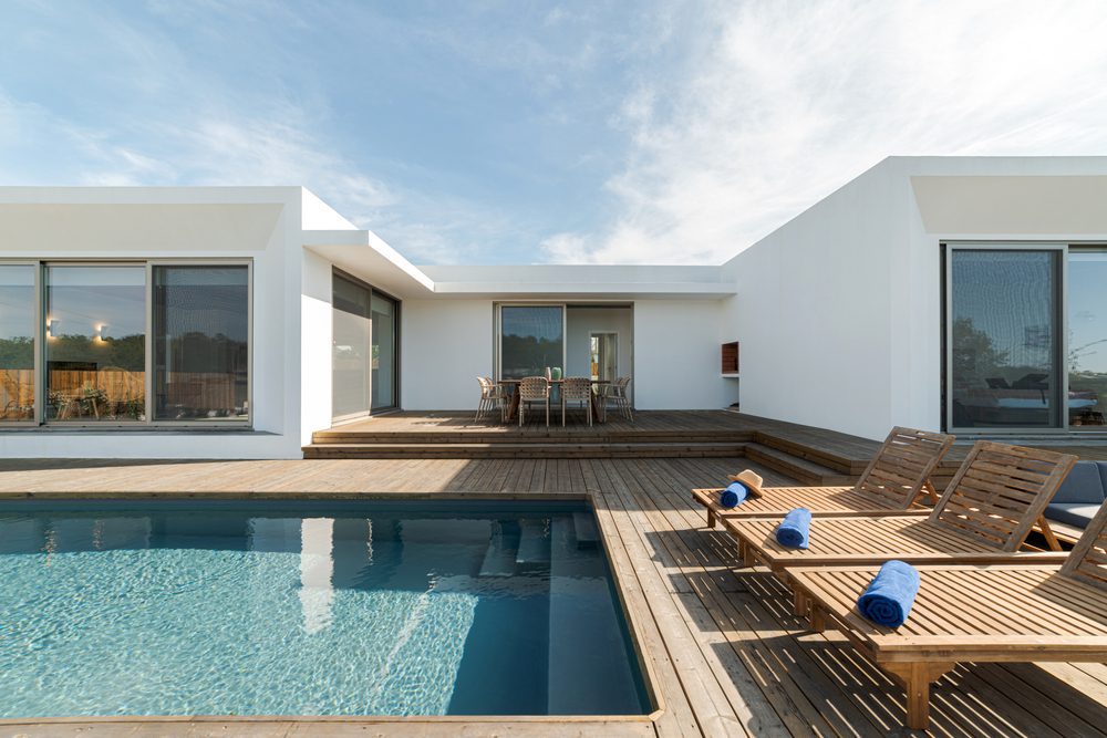 Wooden lounge chairs in modern, contemporary, white villa pool and deck. Blue sky with clouds in the background. Deck chairs with blue rolled up towels beside a built in swimming pool.